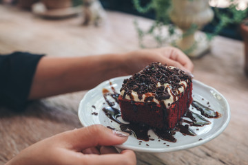A woman holding a piece of red velvet cake with cream and chocolate in white ceramic plate on...
