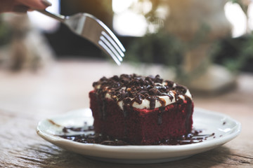 A woman using fork to cut a piece of red velvet cake with cream and chocolate in white ceramic plate on wooden table