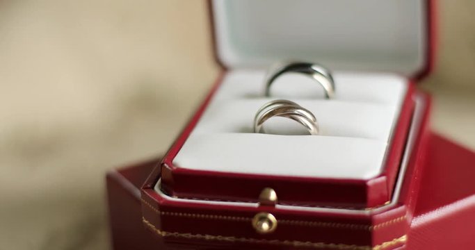 Two designer twisted wedding rings lying in red box on table indoors close up camera sliding. Elegant male female accessories gift for engagement ceremony. Nuptial bridal fashion 4k