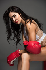 side view of sexy sportive woman in white swimwear and boxing gloves sitting isolated on grey