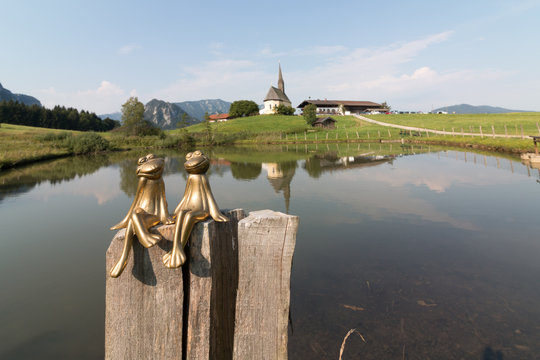 Inzell, Germany - August 5, 2018: View of two frogs in love with the Nikolai Church in Inzell and the Alps in the background.