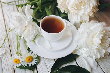 White cup of morning tea and white peonies and chamomile on light gray wooden background. Top view, concept.