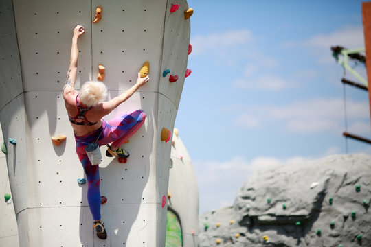 Photo from back of sports blonde with short haircut in sports clothes practicing on wall for climbing against blue sky