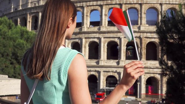 Young woman standing near famous attraction Colosseum in Rome, Italy. Teenage girl waving Italian flag in slow motion. Happy female tourist enjoying european vacation. Student travel through Europe