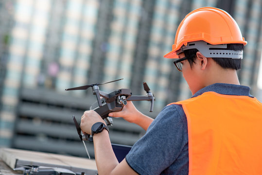 Young Asian engineer adjusting drone at construction site. Using unmanned aerial vehicle (UAV) for land and building site survey in civil engineering project.