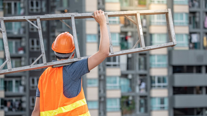 Young Asian maintenance worker man with orange safety helmet and vest carrying aluminium step ladder at construction site. Civil engineering, Architecture builder and building service concepts
