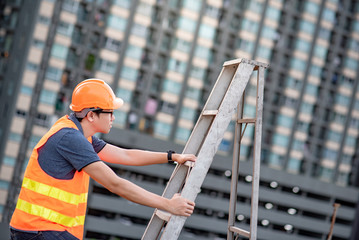 Young Asian maintenance worker man with orange safety helmet and vest carrying aluminium step ladder at construction site. Civil engineering, Architecture builder and building service concepts