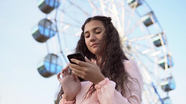 The girl is using a smartphone standing near the Ferris wheel .4K. Close-up