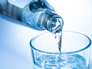 Pouring of cold clear drink water from bottle into glass, close up, blue toned on white background