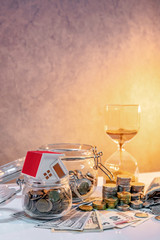 Saving money for retirement concept. Real estate or property investment. Home mortgage loan rate. Coin stack, money bag and currency glass jars on banknotes with hourglasses, house model on the table.