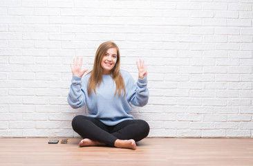 Young adult woman sitting on the floor in autumn over white brick wall showing and pointing up with fingers number nine while smiling confident and happy.