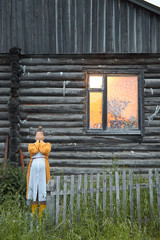 a pregnant woman in a yellow cardigan and a striped dress is standing near an old wooden log house with her hands closed