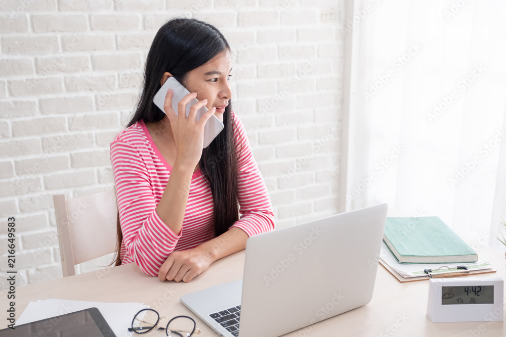 Poster asian female freelancer talk on mobile and see outside window with laptop on table at home.work at h