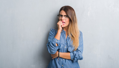 Young adult woman over grunge grey wall wearing glasses looking stressed and nervous with hands on mouth biting nails. Anxiety problem.