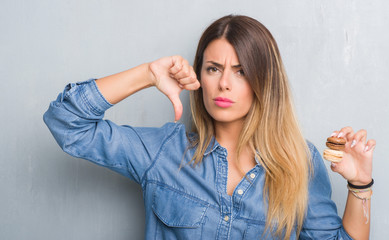Fototapeta premium Young adult woman over grey grunge wall eating chocolate macarons with angry face, negative sign showing dislike with thumbs down, rejection concept