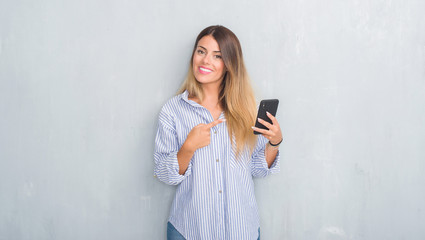 Young adult woman over grey grunge wall looking at smartphone texting a message very happy pointing...