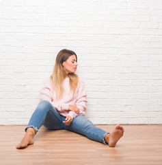 Fototapeta na wymiar Young adult woman sitting on the floor over white brick wall looking to side, relax profile pose with natural face with confident smile.