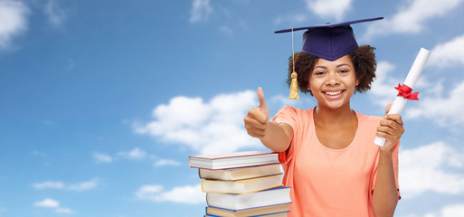 education, school, knowledge and graduation concept - happy smiling african american graduate student girl in mortarboard with books and diploma showing thumbs up over blue sky background