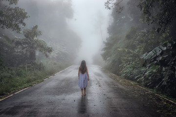 Back view of young woman walking on the road surrounded dense fog and silhouette of tree in winter.