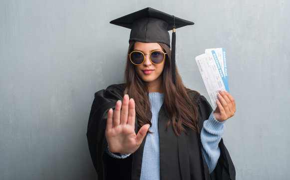 Young brunette woman over grunge grey wall wearing graduate uniform holding boarding pass with open hand doing stop sign with serious and confident expression, defense gesture