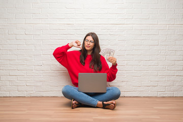 Young brunette woman sitting on the floor over white brick wall paying holding dollars with angry...