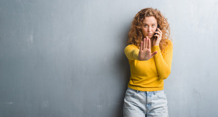 Obraz premium Young redhead woman over grey grunge wall talking on the phone with open hand doing stop sign with serious and confident expression, defense gesture