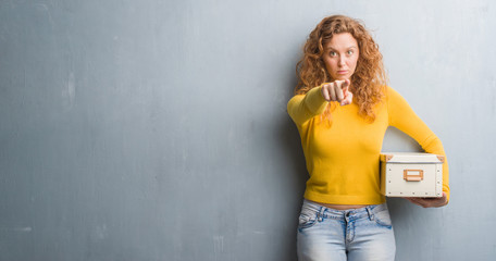 Young redhead woman over grey grunge wall holding box pointing with finger to the camera and to you, hand sign, positive and confident gesture from the front