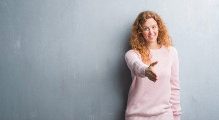 Fototapeta premium Young redhead woman over grey grunge wall wearing pink sweater smiling friendly offering handshake as greeting and welcoming. Successful business.