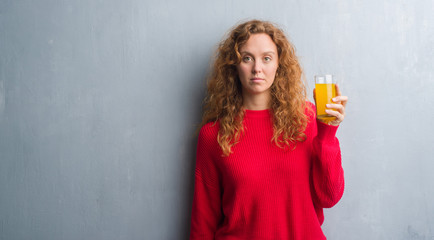 Young redhead woman over grey grunge wall drinking a glass of orange juice with a confident expression on smart face thinking serious