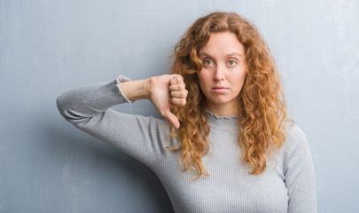 Fototapeta premium Young redhead woman over grey grunge wall with angry face, negative sign showing dislike with thumbs down, rejection concept
