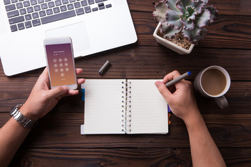 Hand of a man entering password on his smartphone and write something on notebook with wooden desk background