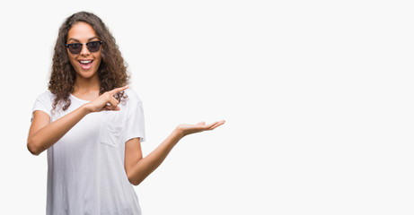 Young hispanic woman wearing sunglasses amazed and smiling to the camera while presenting with hand...