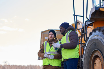 Portrait of two workers, one African-American, standing next to heavy industrial dump truck on...