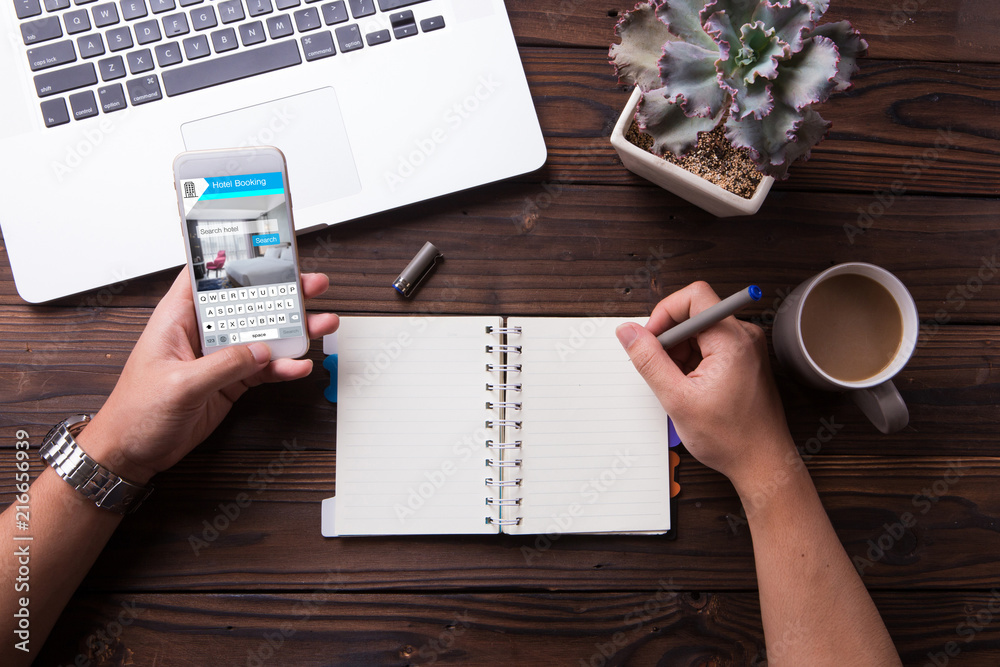 Wall mural man using a smartphone for booking hotel online and write something on notebook with wooden desk bac
