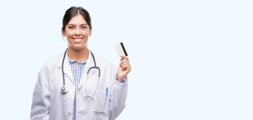 Young hispanic doctor woman holding credit card with a happy face standing and smiling with a confident smile showing teeth