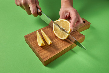 A girls' hands cut yellow organic lemon into slices with knife on a wooden brown board on a green.
