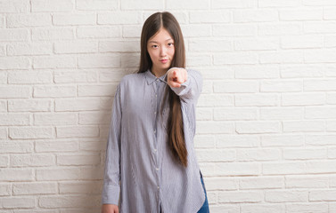Young Chinese woman over brick wall pointing with finger to the camera and to you, hand sign,...
