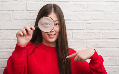 Young Chinese woman over brick wall looking through magnifying glass with surprise face pointing finger to himself
