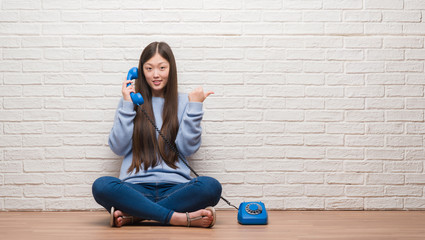 Young Chinese woman calling using vintage telephone pointing and showing with thumb up to the side with happy face smiling