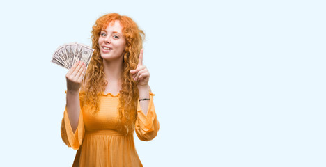 Young redhead woman holding dollars surprised with an idea or question pointing finger with happy face, number one