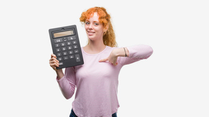 Young redhead woman holding big calculator with surprise face pointing finger to himself