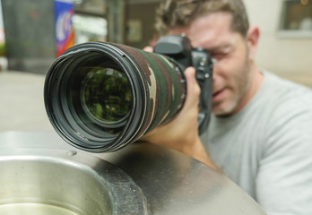 lifestyle funny portrait of young paparazzi photographer man in action hidden behind city paper basket stalking for shooting exclusive photo story