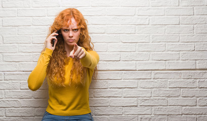 Young redhead woman standing over brick wall talking on the phone pointing with finger to the...