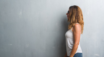 Middle age hispanic woman over grey wall wearing sunglasses looking to side, relax profile pose with natural face with confident smile.