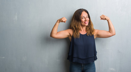Middle age hispanic woman standing over grey grunge wall showing arms muscles smiling proud....