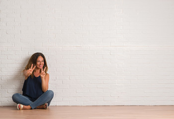 Middle age hispanic woman sitting on the floor over white brick wall smiling looking to the camera showing fingers doing victory sign. Number two.