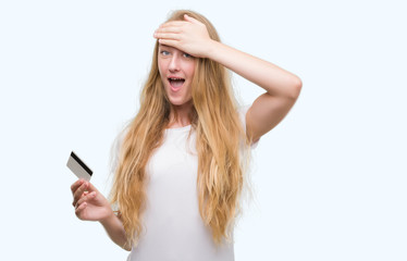 Fototapeta premium Blonde teenager woman holding credit card stressed with hand on head, shocked with shame and surprise face, angry and frustrated. Fear and upset for mistake.