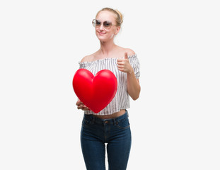Blonde teenager woman holding red heart happy with big smile doing ok sign, thumb up with fingers, excellent sign