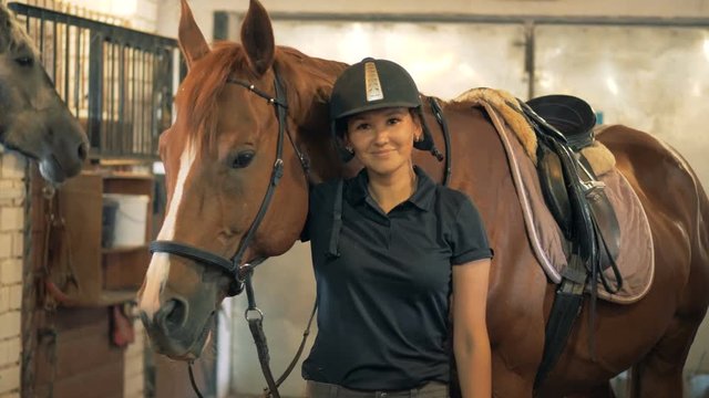 Jockey lady is standing near a brown horse and smiling