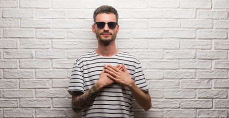 Young adult man wearing sunglasses standing over white brick wall smiling with hands on chest with...